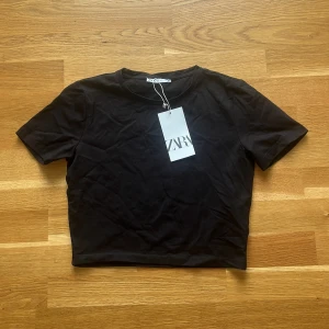 Svart croppad topp från Zara - En svart croppad topp från Zara med rund halsringning och korta ärmar. Enkel och stilren design i mjuk bomull, perfekt att matcha med jeans eller kjol. Snygg basic som funkar till många olika looks. Lappen är kvar!!