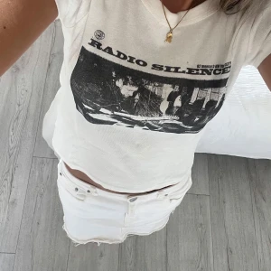 Vit Radio Silence t-shirt från John Galt - Vit t-shirt från John Galt med svart vintageinspirerat tryck på bröstet där det står 'Radio Silence' och en bild på en gitarrist. Modellen är kortärmad och har en klassisk rund halsringning. T-shirten är i mjuk bomull och har en avslappnad, lite croppad passform.