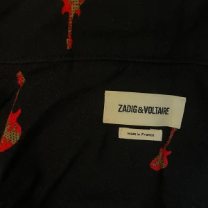 Svart skjorta Zadig & Voltaire med röda detaljer - Svart skjorta från Zadig & Voltaire med röda gitarrmönster och röda passpoaler längs kanterna. Skjortan har knappar framtill, bröstficka och lång ärm. Snygg och unik design som sticker ut.