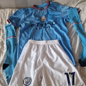Manchester City De Bruyne matchställ - Säljer ett komplett Manchester City matchställ med De Bruyne 17. Tröjan är ljusblå med lång ärm, klubbmärke och Etihad Airways-tryck. Kragen har röda och vita detaljer. Vita shorts med klubbmärke och nummer 17. Ingår även ljusblå strumpor med svarta detaljer. Perfekt för fotbollsfans!