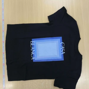 Svart t-shirt från Casablanca - Svart t-shirt från Casablanca med ett stort grafiskt tryck i olika blå nyanser och texten 'CASA BLANCA' på ryggen. Klassisk rund hals och korta ärmar. Perfekt för en stilren och modern look.