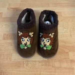 Bruna tofflor med broderad figur - Mysiga bruna tofflor med broderad tecknad figur framtill och mjukt foder inuti. Perfekta för att hålla fötterna varma hemma. Sulan har ett mönster med en nallebjörn.