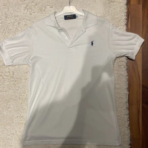 Vit piké från Polo Ralph Lauren - Klassisk vit pikétröja från Polo Ralph Lauren med blå broderad logga på bröstet. Tröjan har korta ärmar, krage och knappar vid halsen. Perfekt för sommaren som snart är här.
