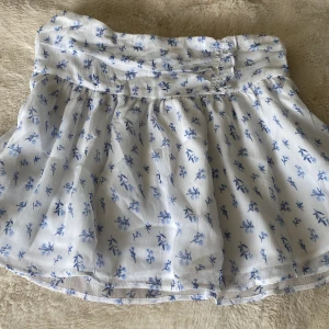 Vit blommig kortkjol från Gina Tricot - Supersöt vit kortkjol från Gina Tricot med blått blommönster. Kjolen är oanvänd oxh säljs inte längre. Säljer då den nu är för liten för mig, och inte används.💕💕