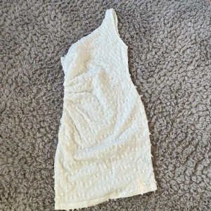 Vit one shoulder klänning - Säljer en vit one shoulder klänning i kort modell. Klänningen har ett diskret strukturerat mönster i tyget och är figurnära. Perfekt för dig som vill ha en stilren och trendig look. Den var tyvärr får stor på mig men super fin och helt ny