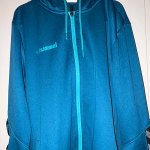Blå zip-hoodie från Hummel - Säljer en blå hoodie från Hummel med dragkedja och huva. Jackan har långa ärmar och en sportig look, perfekt för träning eller chill. Hummel-logga på bröstet och dragsko i huvan.