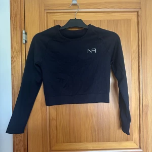 Svart croppad långärmad träningströja från NA - Säljer en svart croppad långärmad träningströja från NA med diskret logga på bröstet. Tröjan har tight passform och är perfekt för gymmet eller andra träningspass.