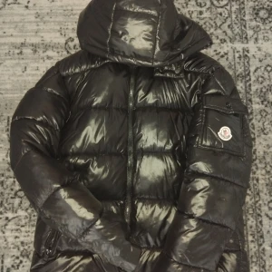 Svart dunjacka från Moncler - Säljer en svart, glansig dunjacka från Moncler med huva och dragkedja framtill. Jackan har en ficka med Moncler-logga på ärmen och quiltad design. Perfekt för kalla dagar och riktigt snygg streetstil.