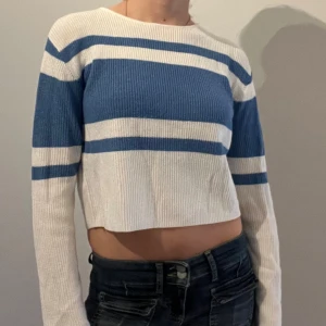 Blåvit randig croppad tröja från H&M - Säljer en croppad långärmad tröja från H&M i ribbat material. Tröjan är vit med breda blå ränder och har en rund halsringning. Perfekt för en avslappnad och trendig stil.
