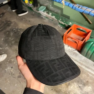 Svart keps från Fendi - Snygg svart keps från Fendi med diskret mönster i tyget och justerbart spänne baktill med Fendi-logga. Klassisk form och stilren design, perfekt för att lyfta din outfit.