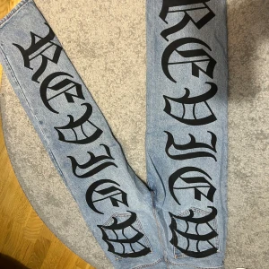 Baggy jeans med tryck från Review - Baggy ljusblå jeans från Review med stora svarta tryck längs båda benen och coolt spindelnätsmotiv vid bensluten. Klassisk femficksmodell med orange sömmar och läderpatch bak i midjan. Perfekt för dig som vill sticka ut.köpta för 1200kr 