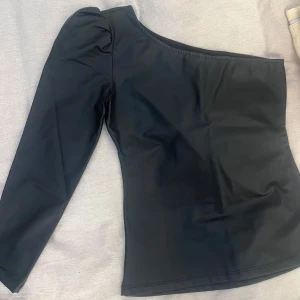 Svart skinaktig one shoulder topp - Snygg svart skinaktig topp med one shoulder-design och lång ärm. Toppen har en stilren look med lätt puff vid axeln och är perfekt för att skapa en trendig outfit. Pris går att diskutera.