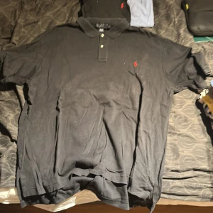 Svart pikétröja från Ralph Lauren - Klassisk svart pikétröja från Ralph Lauren med liten röd broderad logga på bröstet. Tröjan har krage, två knappar och korta ärmar. Perfekt för en stilren och avslappnad look.