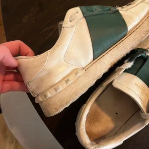 Valentino Open sneakers - Säljer ett par slitna Valentino Open sneakers (innersulan är utbytt) i vitt skinn med grönt band över mitten. Skorna har rund tå, platt sula och klassisk Valentino-detalj baktill. Perfekta för dig som gillar stilrena och exklusiva sneakers.