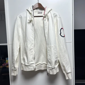 Vit hoodie med dragkedja från Moncler - Säljer en stilren vit hoodie från Moncler med dragkedja framtill och huva. Tröjan har Monclers logga broderad på ärmen. Perfekt för en avslappnad look. Nypris: 9995kr. Köpt på NK Göteborg