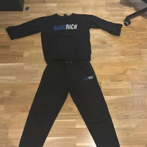 Svart sweatshirt från Hoodrich - Säljer en svart sweatshirt från Hoodrich med tryckt logga i blått och vitt på bröstet. Tröjan har långa ärmar och en klassisk passform. Perfekt för en avslappnad stil. Storlek 13-15 XL (Junior) BYXOR FÖLJER MED!!!