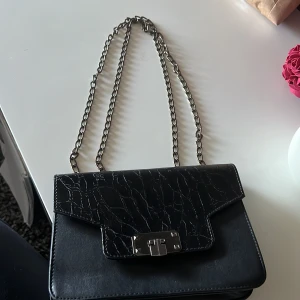Svart axelväska med kedjerem - Säljer en svart axelväska med croco-mönstrad yta och silverfärgad kedjerem. Väskan har ett stilrent metallspänne framtill och fack inuti, inklusive ett med dragkedja. Perfekt för dig som vill ha en snygg accessoar till din outfit.