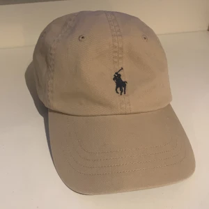 Beige keps från Polo Ralph Lauren - Stilren beige keps från Polo Ralph Lauren med klassisk broderad logga framtill och justerbart spänne bak. Kepsen är tillverkad i bomull och har en avslappnad passform. Helt ny aldrig använd köpt på NK 
