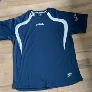 Blå sportig t-shirt från Joma - Säljer en blå t-shirt från Joma med vita detaljer längs axlar och sidor. Perfekt för träning eller fotboll. Tillverkad i syntetmaterial som andas och torkar snabbt. Klassisk passform och kortärmad modell.
