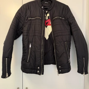Diesel jacka - Snygg svart quiltad jacka från Diesel med flera silverfärgade dragkedjor på bröstet och sidorna. Jackan har lång ärm, ståkrage och coola sömdetaljer. Perfekt för dig som gillar en stilren och modern look.