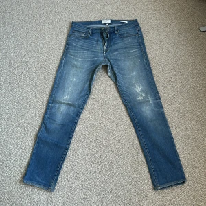 Blå jeans från Frame Denim - Säljer ett par klassiska blå jeans från Frame Denim med lätt slitna detaljer och raka ben. Jeansen har fem fickor, dragkedja och knapp framtill. Perfekta för en avslappnad och stilren look.