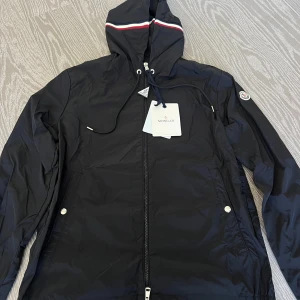 Svart vindjacka från Moncler - Snygg svart vindjacka från Moncler med huva och dragkedja framtill. Jackan har vita detaljer, snörning i huvan och Moncler-logga på ärmen. Perfekt för blåsiga dagar och enkel att matcha med olika outfits.