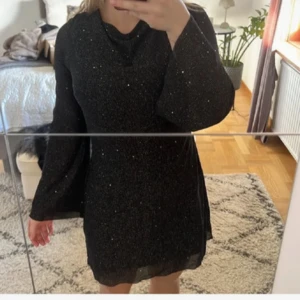 Svart glittrig klänning - Snygg svart glittrig klänning med långa ärmar och en elegant öppen rygg. Perfekt för en kväll ute eller en speciell tillställning. Klänningen har en kort längd och är tillverkad i ett glittrande material som fångar ljuset vackert. Endast Använd 1 gång. 