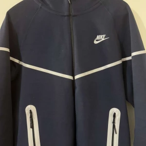 Mörkblå träningsbyxor från Nike - Snygga mörkblå träningsbyxor från Nike med vita detaljer och dragkedjor vid fickorna. Byxorna har smal passform och resår i midjan med snörning. Perfekta för träning eller chill.