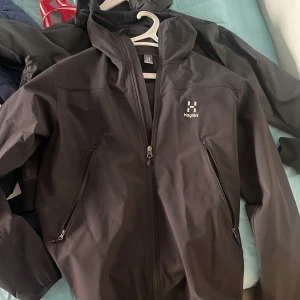 Svart vindjacka från Haglöfs - Säljer en svart vindjacka från Haglöfs med huva och dragkedja framtill. Jackan har två snedställda fickor med dragkedja och en stilren design med logga på bröstet. Perfekt för blåsiga dagar och utomhusaktiviteter.