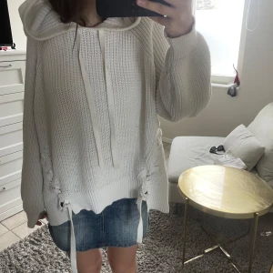 Vit oversized stickad hoodie med snörning - Säljer en vit, oversized stickad hoodie med huva och långa ärmar. Tröjan har snörningsdetaljer vid nederkanten och grov ribbstickning. Perfekt för mysiga dagar. 