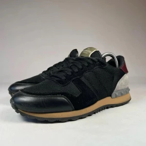 Valentino Rockrunner  - Snygga Valentino Rockrunner sneakers i svart   skinn med grå och vinröda detaljer. Skorna har en beige mellansula och grå yttersula med nitar vid hälen. Perfekta för dig som vill ha en stilren och exklusiv sneaker.