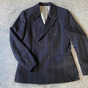 Mörkblå randig kavaj från Tiger of Sweden - Dubbelknäppt mörkblå kavaj med diskreta ränder från Tiger of Sweden. Klassisk passform med fyra knappar framtill och tre knappar vid ärmslut. Fodrad insida och fickor framtill. Perfekt för en stilren look.