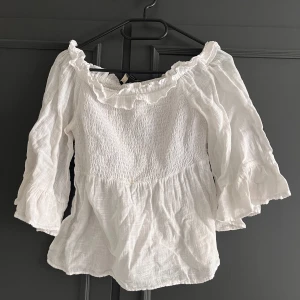 Vit offshoulder blus med volang - Superfin vit blus med offshoulder och volangdetaljer. Har trekvartsarmar. Är köpt secondhand och har en liten fläck, men som knappt syns, därför priset.