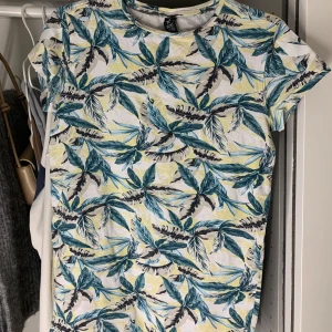 Mönstrad t-shirt från SOC - Säljer en fräsch t-shirt från SOC med tropiskt blad-mönster i blått, grönt och gult på vit botten. T-shirten har korta ärmar och är perfekt för sommardagar. Skön och lätt att bära.