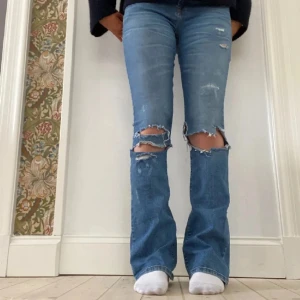 Blå slitna jeans - Säljer ett par blå jeans med slitningar på knäna. De har en bootcut-stil och är lågmidjade. Perfekta för en avslappnad look.