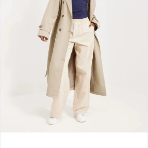 Beige trenchcoat med bälte - Snygg beige trenchcoat med klassisk krage och knappar framtill. Jackan har ett matchande bälte i midjan och långa ärmar med knappar vid ärmslut. Perfekt för lager-på-lager och stilrena outfits.