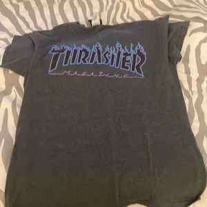 Grå Thrasher t-shirt med blå och lila tryck - Säljer en grå t-shirt från Thrasher med ikoniskt blått och lila flamtryck på bröstet. Klassisk passform och rund halsringning. Perfekt för dig som gillar streetwear och skatekultur. Exklusiv och går inte att köpa längre