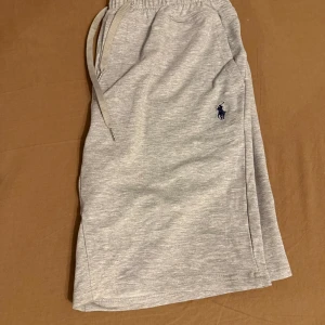Ljusgrå shorts från Polo Ralph Lauren - Snygga ljusgrå shorts från Polo Ralph Lauren med klassisk broderad logga i mörkblått på ena benet. Shortsen har snörning i midjan och är tillverkade i mjuk bomull, perfekta för en avslappnad stil.