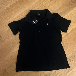 Svart pikétröja från Peak Performance - Klassisk svart pikétröja från Peak Performance med liten vit logga på bröstet. Tröjan har krage och knäppning framtill samt korta ärmar. Perfekt för en sportig och stilren look. I storlek M