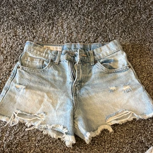 Ljusblå jeansshorts från Zara - Säljer ett par ljusblå jeansshorts från Zara med fransade benslut och slitna detaljer. Klassisk femficksmodell med dragkedja och knapp framtill. Perfekta för sommaren och riktigt snygga till sneakers eller sandaler.