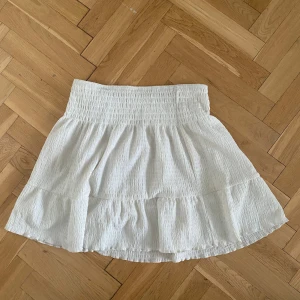 Vit volangkjol med resårmidja - Säljer en söt vit kjol med volang och smockad resår i midjan. Kjolen är kort och har en luftig känsla, perfekt för varma dagar. Materialet är lätt och har en krinklad struktur som ger en avslappnad look.