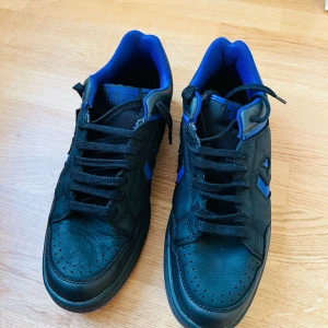 Svarta Converse sneakers med blå detaljer - Säljer ett par svarta Converse sneakers med blå detaljer och svart snörning. Skorna har låg modell, rund tå och platt sula. Perfekta för dig som gillar klassisk streetstil med en twist av färg.