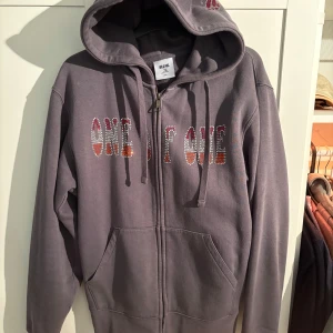 Lila hoodie med strasstenar från One of One - Säljer en lila hoodie från One of One med dragkedja och huva. Framsidan har text i strasstenar och på huvan finns en fjäril i samma stil. Mjuk insida och två fickor  framtill. Perfekt för dig som gillar unika detaljer!