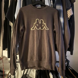 Brun sweatshirt från Kappa - Mörkbrun sweatshirt från Kappa med det klassiska logotyptrycket i vitt på bröstet. Tröjan har rund halsringning och långa ärmar. Perfekt för en avslappnad och sportig stil.