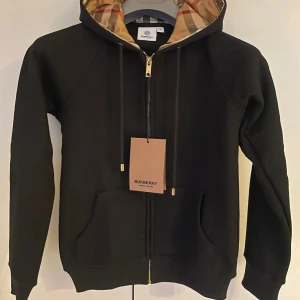 Svart hoodie från Burberry med klassiskt rutigt foder - Säljer en svart hoodie från Burberry med dragkedja och klassiskt rutigt foder i huvan. Tröjan har guldiga detaljer på dragkedjan och snörändarna samt två fickor framtill. Perfekt för dig som gillar lyxiga streetwear-plagg.