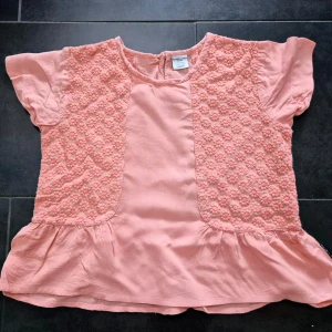 Rosa blus med blommiga detaljer från Polarn O. Pyret - Söt ljusrosa blus från Polarn O. Pyret med korta ärmar och volang nertill. Framsidan har broderade blommor och en liten knapp i nacken. Perfekt för vår och sommar! Ny aldrig använd 