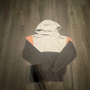 Grå och svart hoodie med rosa detaljer - Säljer en hoodie i grått och svart med rosa detaljer på axlarna. Tröjan har huva och långa ärmar, perfekt för en avslappnad stil. Passar dig som gillar färgblockade plagg.