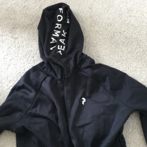 Svart hoodiejacka från Peak Performance - Svart hoodiejacka från Peak Performance med dragkedja och vit logga på bröstet. Huva med tryckt text på insidan. Perfekt för träning eller chill. Snygg och enkel design med långa ärmar och fickor med dragkedja.