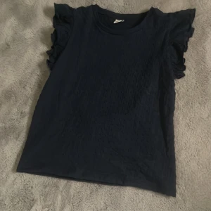 Mörkblå topp med volangärmar från Lindex - Säljer en mörkblå topp från Lindex med fina volangdetaljer på ärmarna. Toppen har rund halsringning och är perfekt för dig som gillar en enkel men ändå söt stil. Säljer då jag inte tycker jag passar i den!😊💕