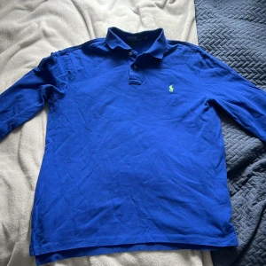 Blå långärmad pikétröja från Polo Ralph Lauren - Klassisk blå långärmad pikétröja från Polo Ralph Lauren med den ikoniska gröna loggan broderad på bröstet. Tröjan har krage och knäppning framtill. Perfekt för en stilren och avslappnad look.